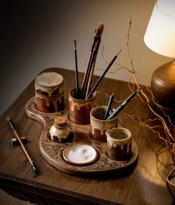 Handcrafted ceramic set for painting and calligraphy on stylish wooden seat. Brushes, calligraphy pens, palette, inks on wooden table with warm table lamp light. Artist, art and hobby.